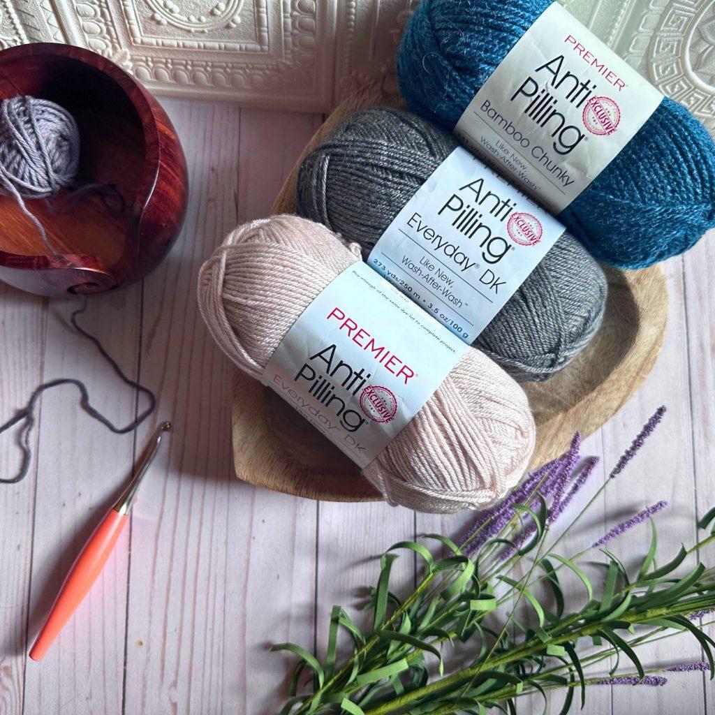three skeins of anti pilling yarn in shades of beige, gray and teal. there is a crochet hook and sprig of lavender flowers nearby