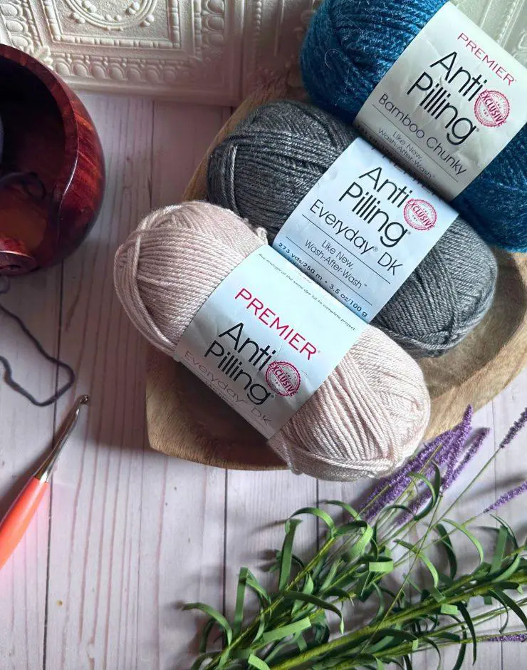 three skeins of anti pilling yarn in shades of beige, gray and teal. there is a crochet hook and sprig of lavender flowers nearby