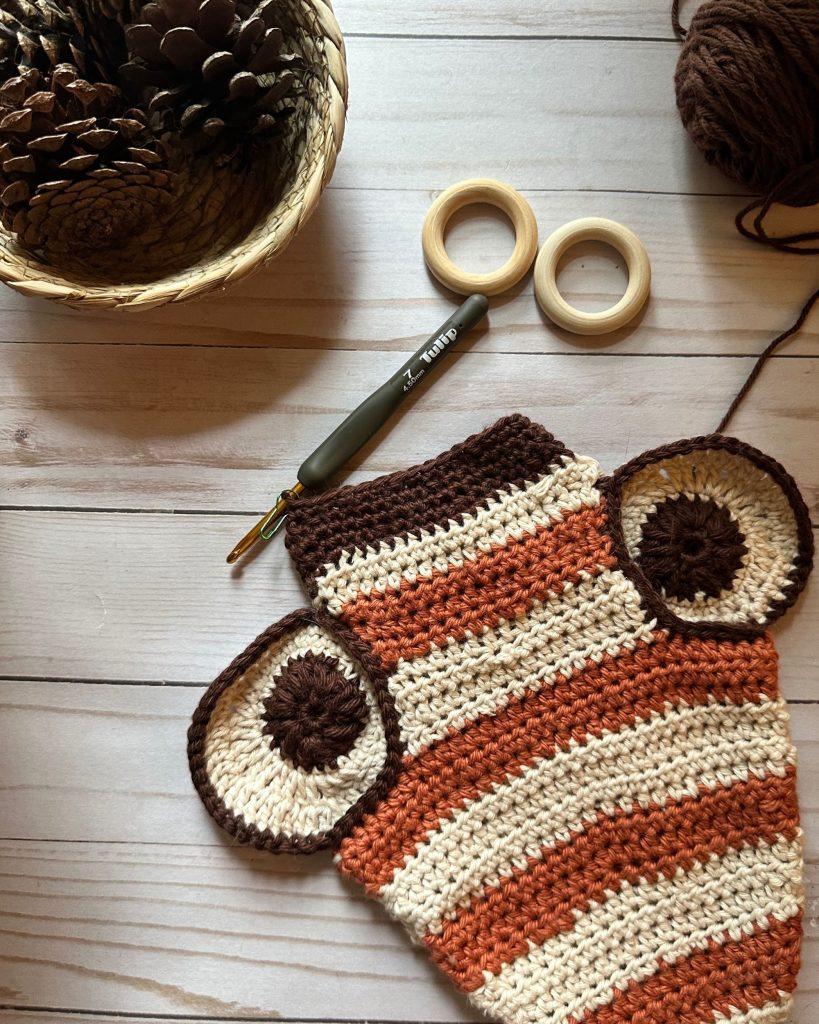 crochet bee potholder in progress. it is whimsical made in fall yarn colors, cream, rust and deep brown, on a white wood table with a baske of pinecones nearby