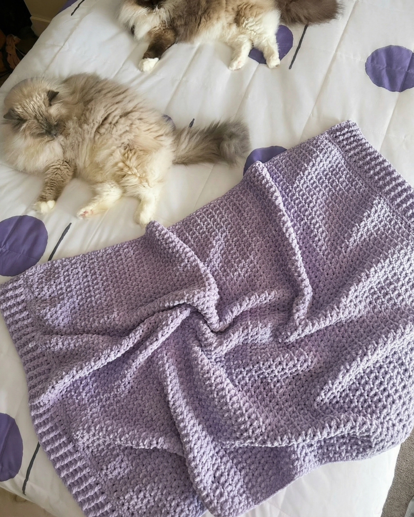 a chunky chenille throw crocheted in a lavender yarn on a bed with two fluffy cats